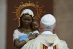 Statue of Mary holding infant Jesus with a gold halo, as a priest in cream robes faces away.