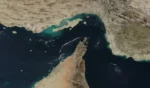 Satellite image of a desert coastline: a sandy peninsula extends into dark blue sea with turquoise shallow waters near the shore.
