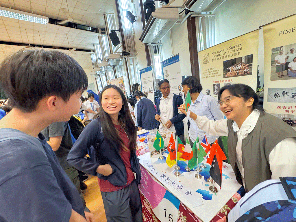 Students chat at a cultural fair table displaying multiple country flags and informational banners in the background, smiling and engaging with attendees.