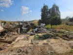 Pile of rubble and collapsed building debris in a rural yard under a bright blue sky, with two trees in the background and scattered scraps around. (informative)