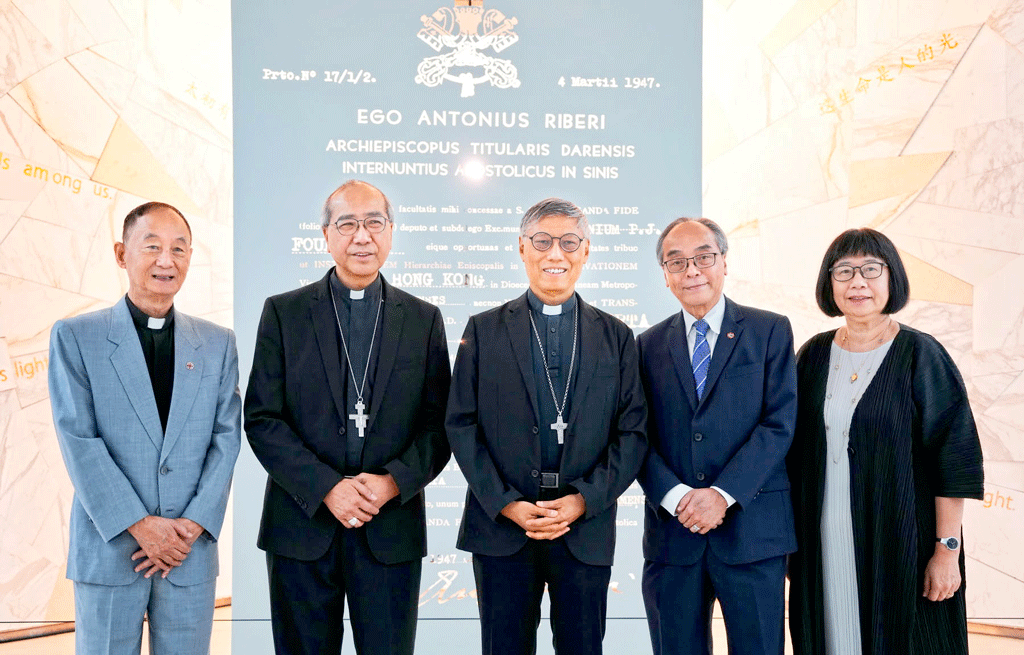 Five people pose for a formal indoor group photo; two clergy in black robes with crosses stand with three professionals in front of a blue inscription panel.
