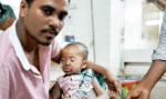 Man cradling a frail baby with an oxygen mask in a clinic setting