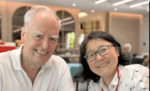 Older man with white hair in a white polo shirt and a smiling woman with glasses in a bright cafe, posing for a selfie on a casual outing.
