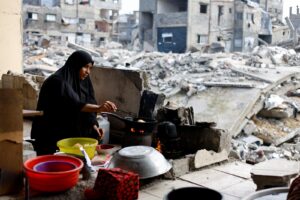 Woman in a black hijab cooks over a small stove amid rubble and ruined buildings behind her.