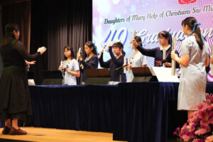 Group of students on a stage holding candles, led by a conductor on the left; banner behind them reads 'Daughters of Mary Help of Christians.'