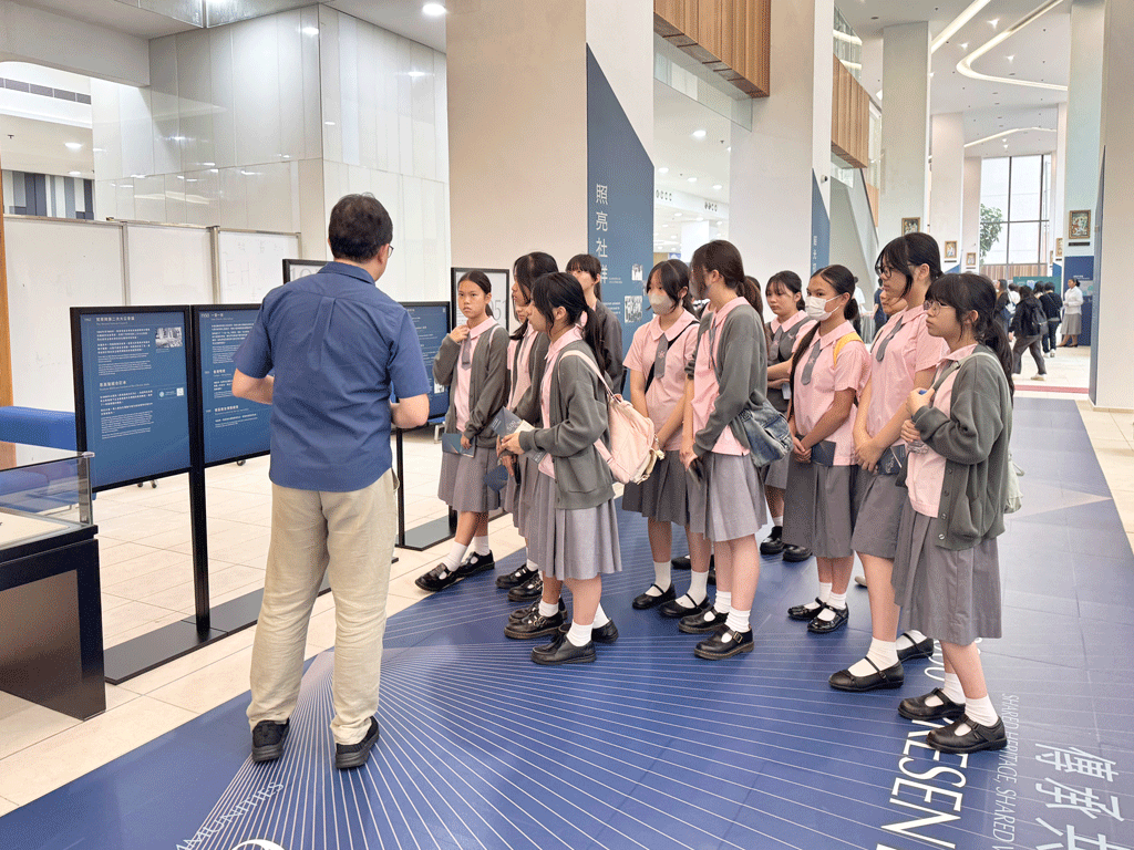 A tour guide explains exhibit panels to a line of uniformed students in a gallery filling with blue information boards.