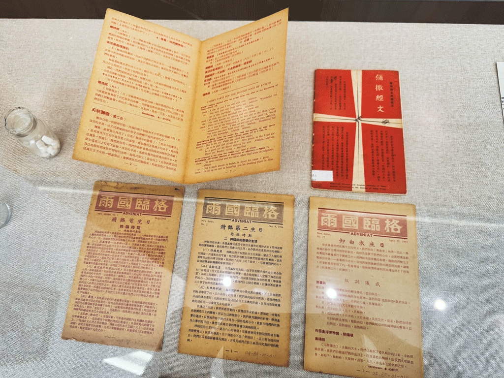 Open, yellowed book and four Chinese pamphlets arranged on a glass display case with a small salt shaker nearby.
