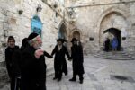 Group of Orthodox Jewish men in black coats holding hands in a stone courtyard with arches and blue doors in the background