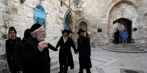 Group of Orthodox Jewish men in black coats holding hands in a stone courtyard with arches and blue doors in the background