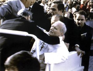 An elderly man in white robes being assisted into a vehicle by security guards as a crowd looks on.
