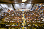 Crowded church interior with worshippers seated in pews facing the altar and a crucifix mounted on the front wall.