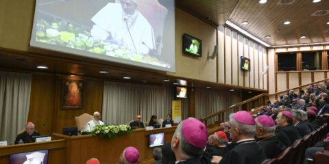 Panel of bishops and clergy seated at a long conference desk in a large meeting hall, with a projection screen showing a speaker in white robes and floral decor at the front.