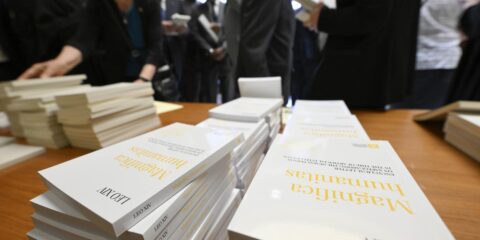Stacks of white books on a wooden table at a crowded event, attendees browsing in the background.