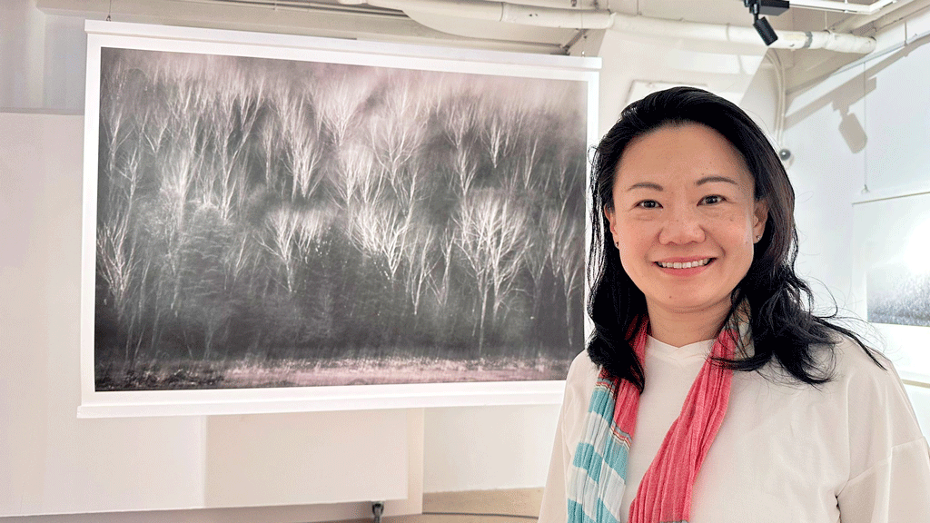 Smiling woman standing next to a large framed black-and-white photograph of a forest in a white-walled gallery room.