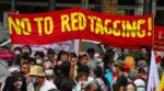 Crowd of protesters wearing masks under a red banner that reads 'NO TO RED TAGGING!'