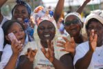 Joyful group of African women and girls at an outdoor event, raising hands and smiling, wearing white shirts and headbands.