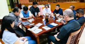 A diverse group of professionals sits around a wooden conference table, discussing documents and tablets in a formal meeting.