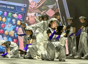Group of performers in shiny silver outfits with purple gloves kneeling and using tablets on a stage, with a giant smartphone game screen backdrop behind them.