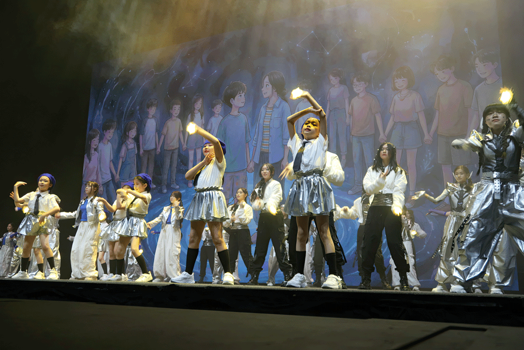Group of young performers dancing on a stage in silver outfits, hands raised, with a painted backdrop behind them.