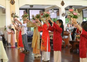 Group of people in colorful áo dài holding bouquets aloft during a ceremony in a hall.