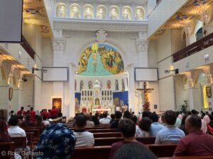 Interior of a church filled with parishioners facing the altar, crucifix to the right, ornate façade and stained-glass windows behind the altar illustrative scene central to the service.