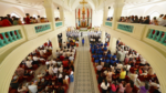 Interior of a large church filled with worshippers, a central aisle leads to an ornate altar with a choir in blue shirts at the front.