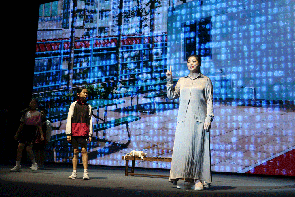 Woman in a long light-gray dress on stage points upward as two children stand nearby, with a digital cityscape and binary-like backdrop behind them.