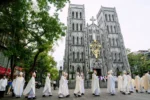 Group of Catholic clergy in white robes walk in a procession past a gothic grey stone church with twin towers and a gold cross above the entrance, on a city street.