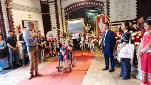 Elderly woman in a wheelchair on a red carpet, speaking to a man with a microphone during a formal ceremony with attendees and floral displays behind them.
