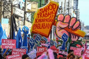 Protest crowd holding pink signs and a large orange sign with red Filipino text, and a giant pink raised-fist prop in a busy street.