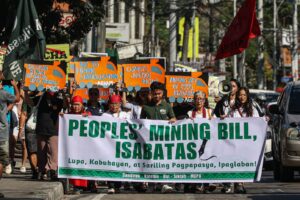 Group of protesters marching on a street holding a large banner that reads 'PEOPLES' MINING BILL, ISABATAS' with smaller text below in a Filipino dialect.