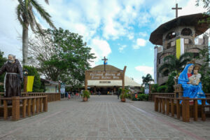 Front view of the National Shrine of Saint Padre Pio entrance, with a wooden sign, cross, and statues on both sides in a sunny plaza.