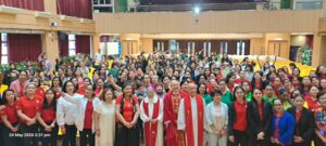 Group photo in a hall with many people, clergy in red vestments at center and others around; timestamp 24 May 2026 5:31 pm visible in corner.