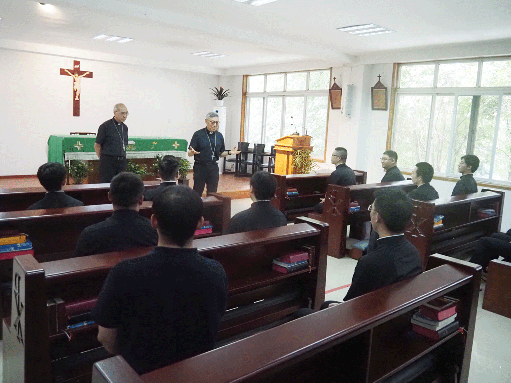 Priests address a group of men seated in wooden pews inside a chapel. A crucifix hangs on the wall behind them and a green altar cloth covers the table at the front.