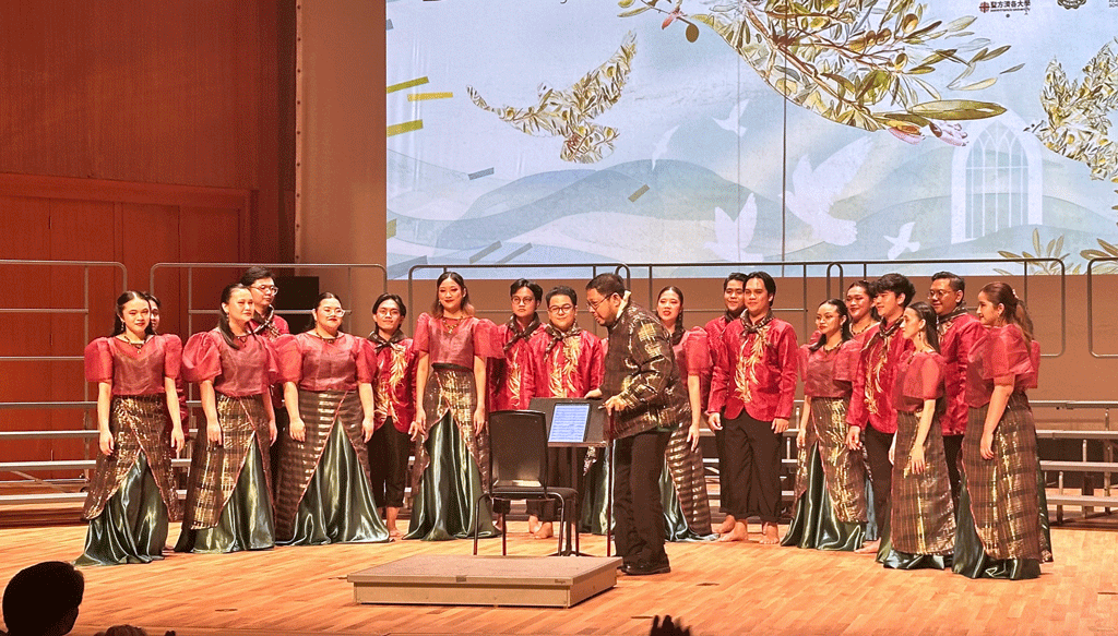 Chorus of students in red blouses and green plaid skirts perform on a wooden stage, conductor guiding them in front of a backdrop mural.