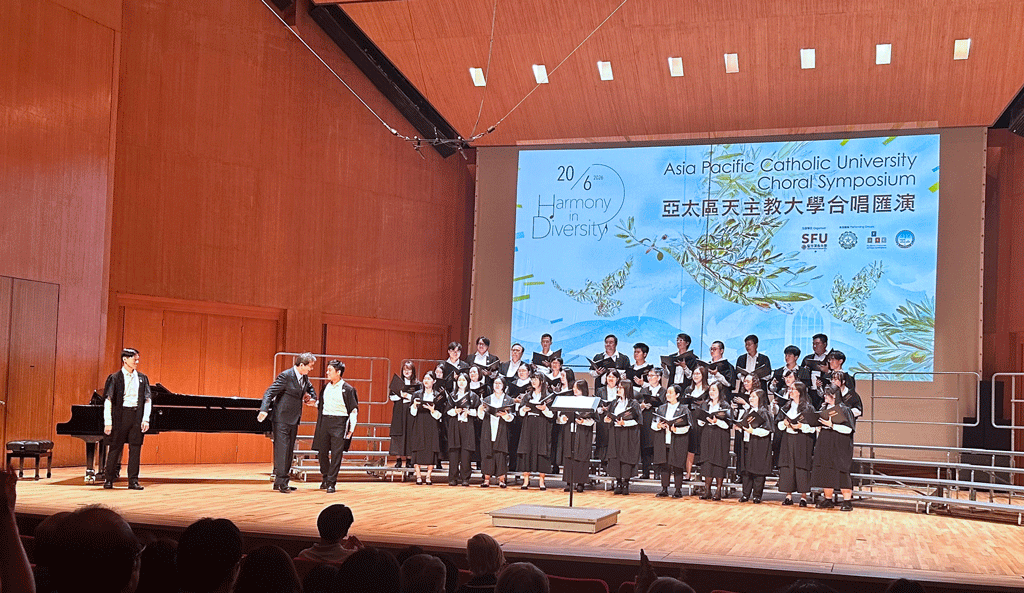 Choir in black robes performs on a wood-paneled stage with a conductor and pianist, under a banner for the Asia Pacific Catholic University Choral Symposium and Harmony in Diversity.