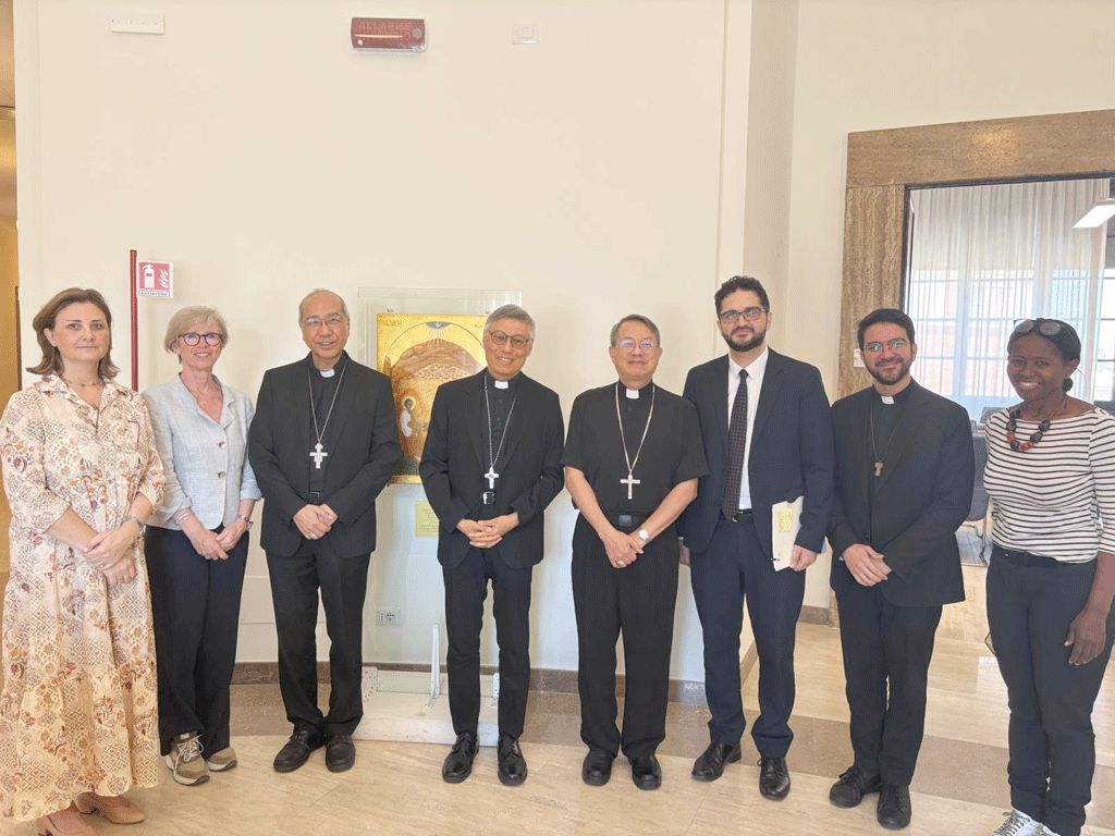 Eight people stand in a bright hallway for a group photo, four priests with crosses and civilians, with a framed religious icon in the background on a cabinet.