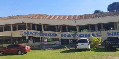 Two-story beige building with a rusted metal roof behind a chain-link fence; large blue letters spell 'NATIONAL' on the front, with red and white cars parked on the grass in front of it.