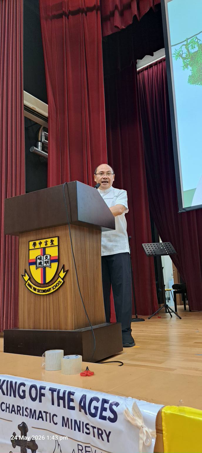 Speaker at a wooden podium on a stage, delivering a talk with a school crest on the front and a banner reading 'King of the Ages Charismatic Ministry'