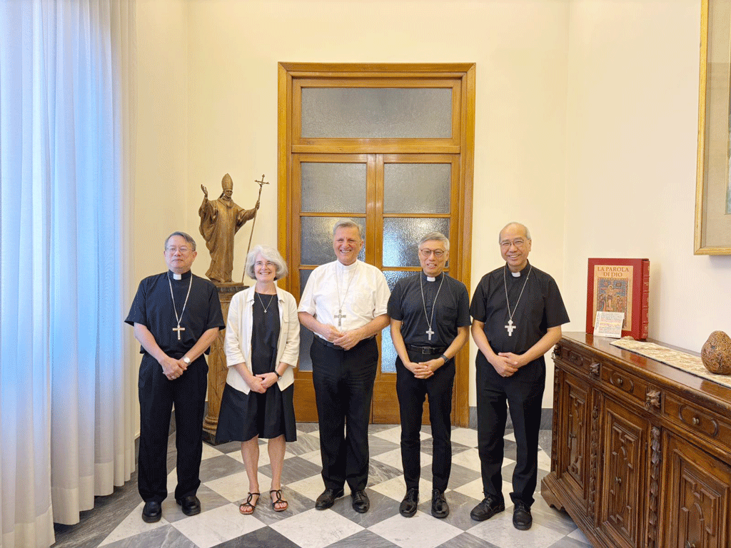 Five people stand in a church-like hallway, four men in black clerical clothes and a woman in a light jacket, with a statue and wooden cabinet behind them.