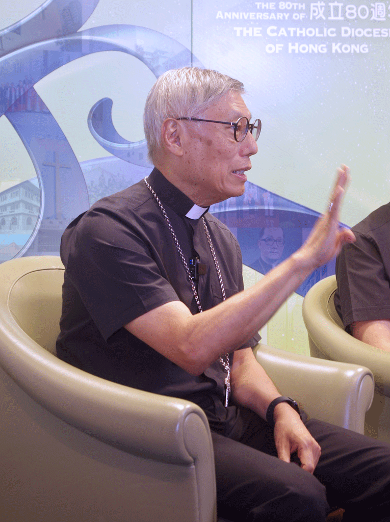 Catholic priest with glasses and a clerical collar sits in a beige chair and gestures with his right hand during a panel discussion.