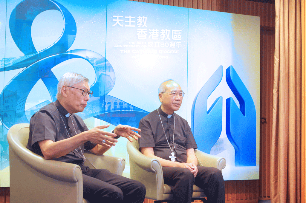 Two clergy members with crosses sit in beige armchairs on a stage, discussing on a blue-themed backdrop backdrop with abstract graphics.