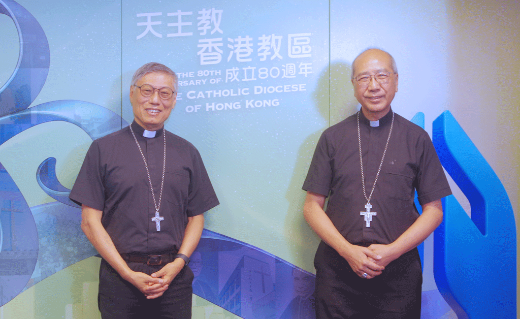 Two Catholic priests in clerical collars and cross necklaces stand side by side in front of a blue-green backdrop with Chinese characters and 'Catholic Diocese of Hong Kong'.