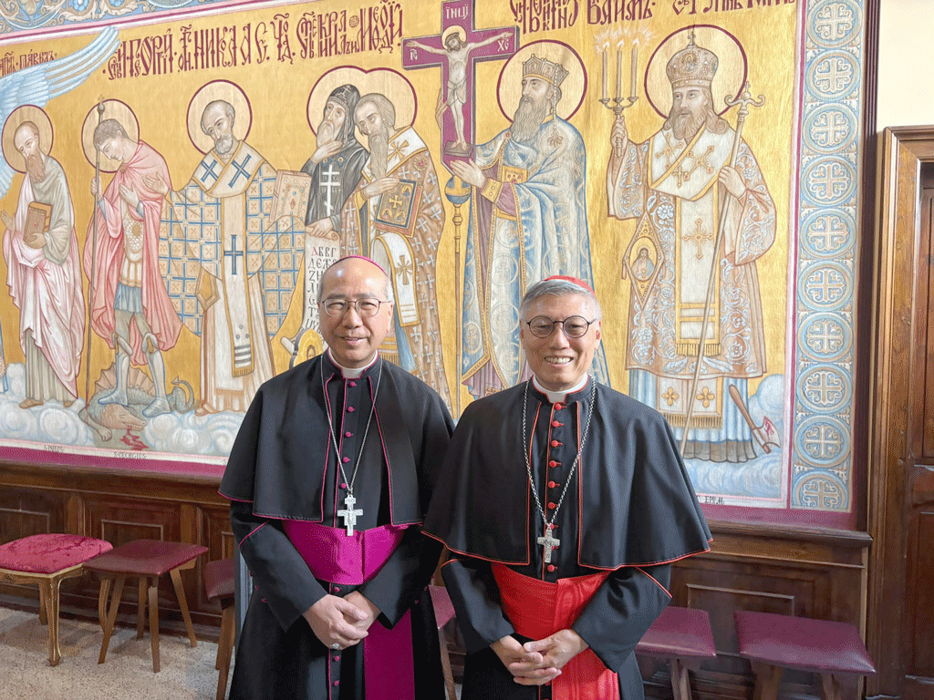 Two Catholic bishops in black vestments with magenta and red sashes stand in front of a colorful religious mural.