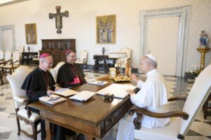 Pope in white robes sits at a wooden desk across from two bishops wearing black and red attire in an ornate room.