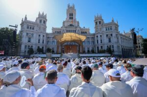 Large crowd in white robes attending an outdoor ceremony in front of a grand white neoclassical building with a stage and Spanish flag.