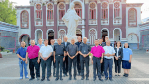 Group of priests and staff posing outside a pink church with a statue in front.