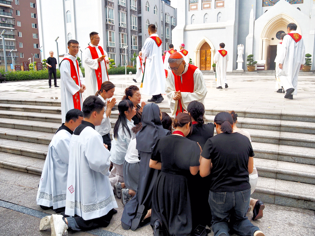 Parishioners kneel in prayer before a bishop and priests in white and red robes outside a church.