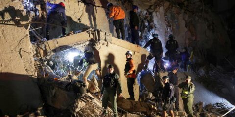 Rescue workers and soldiers on a shattered concrete wall at night, navigating rubble with ropes and flashlights during a search operation.