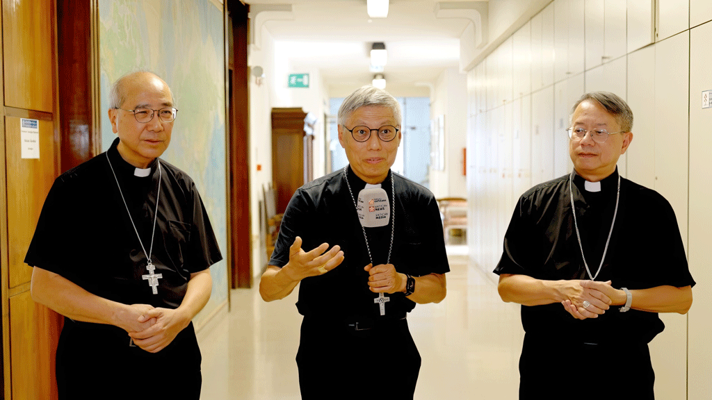 Three Catholic priests in black clerical shirts stand in a hallway, the center priest speaking into a microphone.